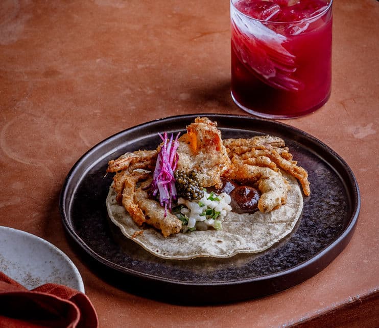 A close-up of a prepared taco on a black plate with fried soft shell crab, garnishes, and a red cocktail drink in the background.