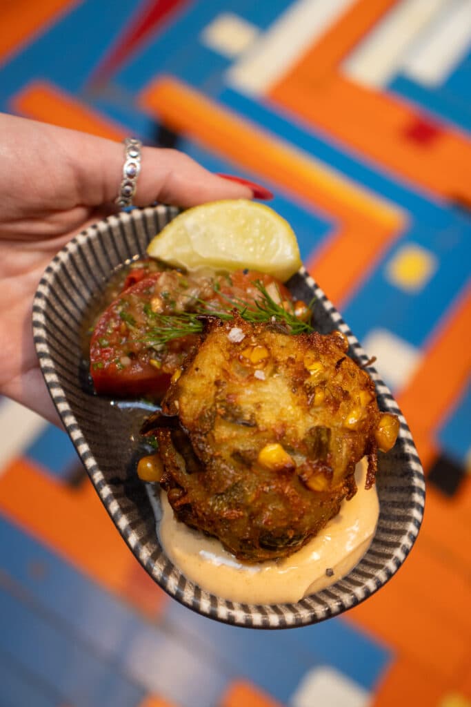 Hand holding a dish with a savory fritter topped with corn, a lemon wedge, tomato salsa, and a creamy sauce.
