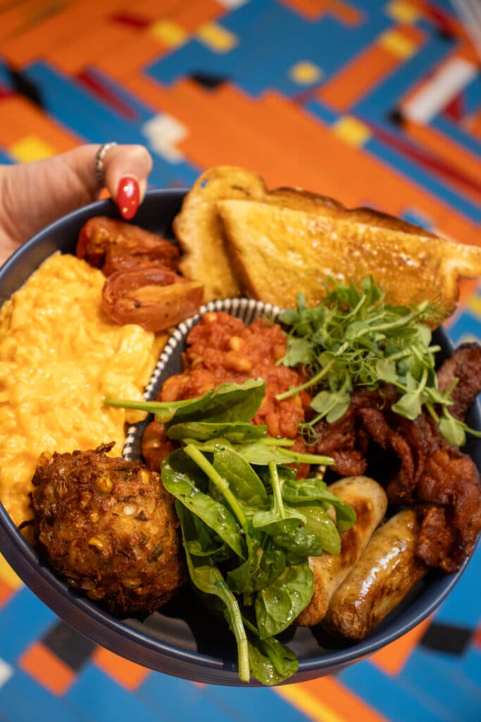 A close-up of a breakfast plate featuring scrambled eggs, toast, sausages, bacon, greens, and a fried item.