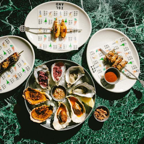 Plates of skewered meats and a platter of oysters served with various sauces on a green marble table.