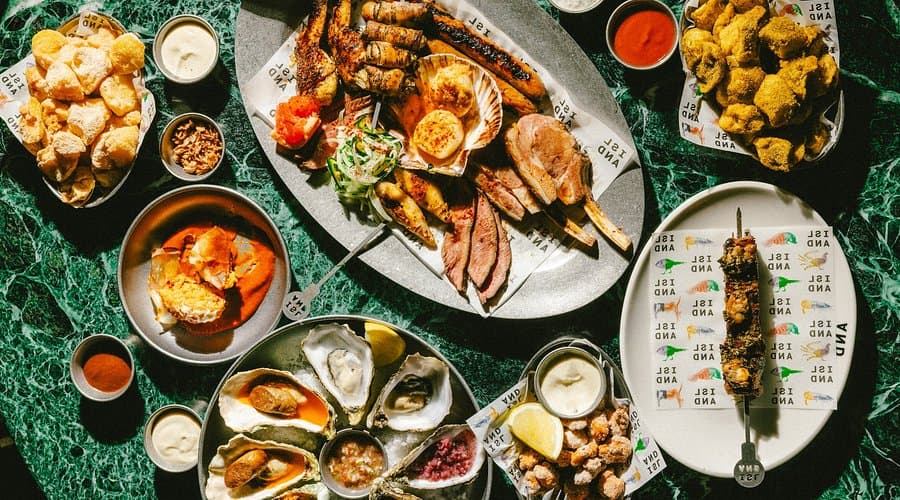 A variety of seafood and grilled meats served on plates and a platter, accompanied by sauces, on a green marble table.
